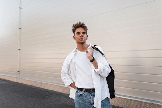 Europäischer junger Mann mit einer stilvollen Frisur in einem trendigen weißen Hemd in Vintage-Jeans steht in der Nähe eines modernen weißen Gebäudes