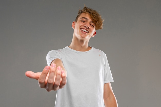 Europäischer lächelnder blonder Kerl in einem weißen T-Shirt ruft mit seiner Hand zu sich selbst an einer grauen Wand
