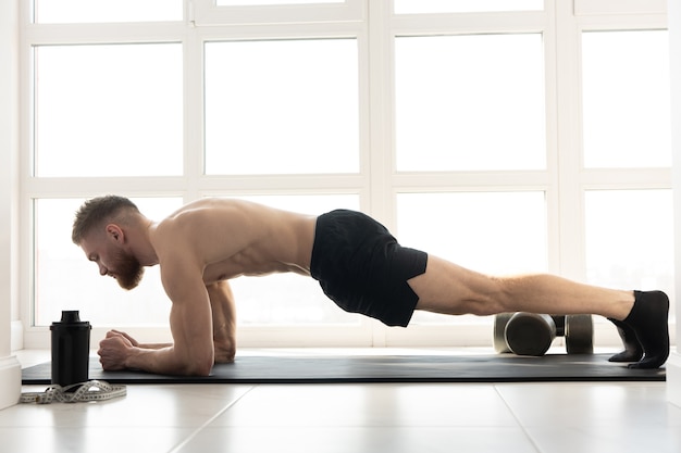 Europäischer Sportler, der Planke für Bauchmuskeln auf Fitnessmatte macht. Junger bärtiger Mann mit nacktem Oberkörper. Konzept der sportlichen Aktivität zu Hause. Innenraum der modernen geräumigen Wohnung. Sonniger Tag