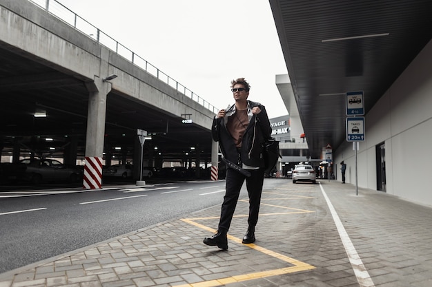 Europäischer trendiger junger Hipster-Mann in modischer Jugendschwarzkleidung in Vintage-Sonnenbrillen mit einem stylischen Rucksack reist an einem Frühlingstag auf der Straße. Urbaner Kerl im Freien. Kleidung und Accessoires für Männer.