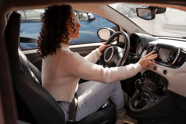 Europäisches Mädchen, das Bildschirm im Auto drückt. Fokussierte junge lockige Frau mit Brille. Moderne Frau als Fahrerin im Luxusautomobil. Konzept des Autofahrens