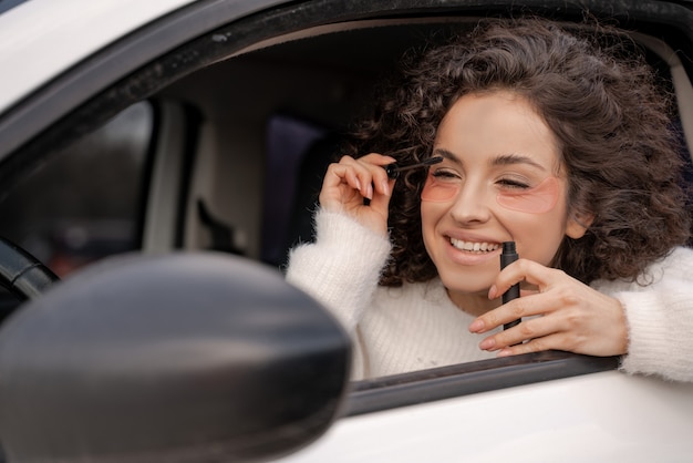 Europäisches Mädchen im Auto trägt Wimperntusche mit Augenbürste auf die Wimpern auf. Lächelnde lockige Frau mit Augenklappe im Gesicht, die den Autospiegel betrachtet. Multitasking. Moderner Lebensrhythmus. Konzept der Gesichtspflege