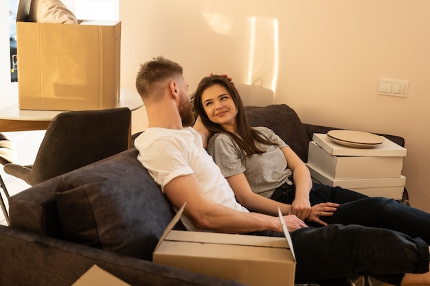 Europäisches Paar sitzt und ruht sich zu Hause auf dem Sofa aus. Mann, der seine lächelnde Freundin streichelt. Konzept des Einzugs in eine neue Wohnung. Idee einer jungen Familie. Kartons mit Sachen. Interieur des Studio-Apartments