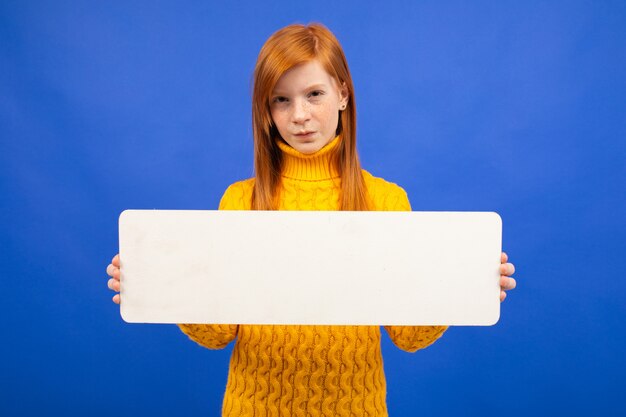 Foto europäisches rothaariges junges mädchen, das ein weißes blatt papier mit einem modell für werbung auf einem blauen studiohintergrund hält