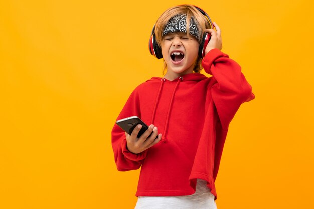Europeo sonriente niño rubio en una sudadera con capucha roja concentrarse en escuchar música en auriculares rojos y tiene teléfono inteligente en amarillo