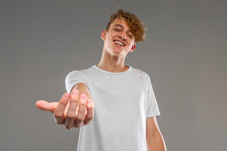 Europeu loiro sorridente em uma camiseta branca está chamando com a mão para si mesmo em uma parede cinza