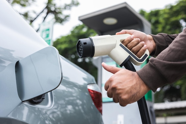 Ev car Coche eléctrico cargando en el estacionamiento. Mano masculina que muestra los pulgares hacia arriba sosteniendo el cable de alimentación conectado a la estación de carga de automóviles eléctricos. Energía alternativa ecológica. Futuro vehículo a batería.