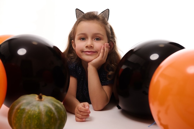 Evento tradicional, conceito de festa de Halloween. A bruxinha linda no aro com orelhas de gato deitada no fundo branco com espaço de cópia ao lado de balões pretos laranja e abóbora