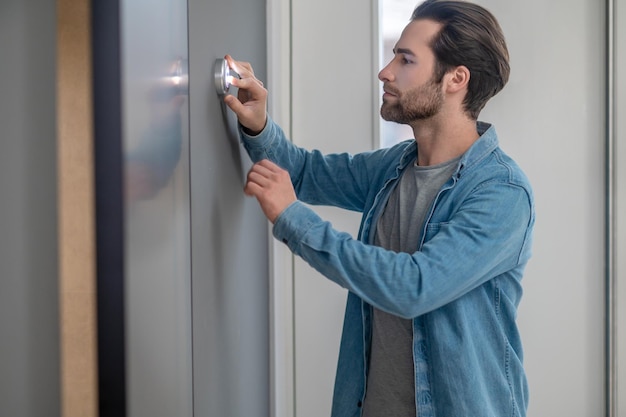 Examen. Joven barbudo de pie de lado a la cámara mirando atentamente tocando el dispositivo en la pared interior