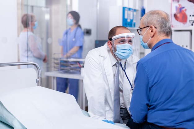 Examen del latido cardíaco del paciente con estetoscopio en la habitación del hospital y uso de mascarilla como precaución de seguridad contra el coronavirus