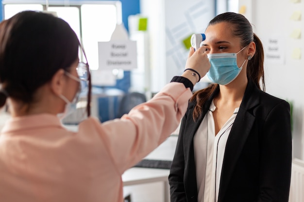 Examinando a temperatura corporal de um trabalhador de escritório com termômetro digital durante a pandemia global com coronavírus. Mulher usando máscara facial como prevenção da infecção com covid19. Nova empresa normal.