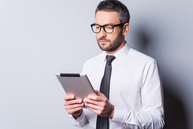 Examinando una nueva tableta. Hombre maduro confiado en camisa y corbata sosteniendo tableta digital y mirándola mientras está de pie contra el fondo gris