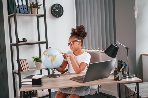 Examinando o globo Uma linda estudante afro-americana está sentada ao lado do laptop