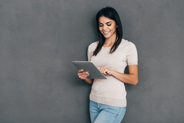 Examinando seu novo tablet. Mulher jovem e atraente em roupa casual trabalhando em um tablet digital e sorrindo em pé contra um fundo cinza