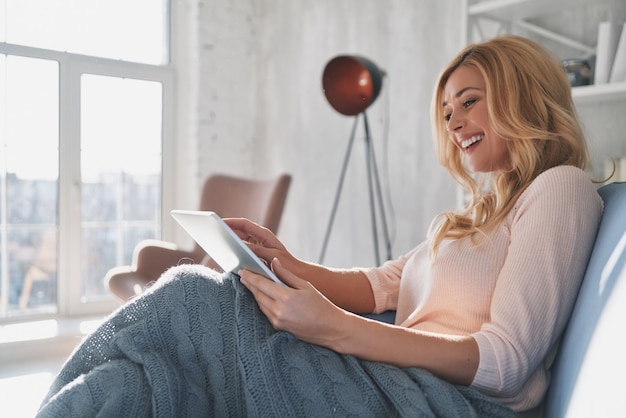 Examinando su nueva tableta. Mujer joven atractiva con tableta digital y sonriendo mientras está sentado en el sofá en casa