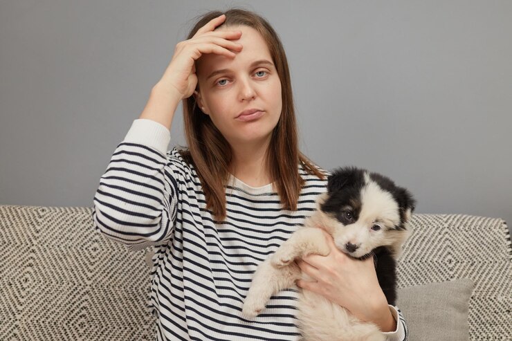 Exausta mulher caucasiana vestindo camisa listrada sentada no sofá com um cão pequeno sendo pneu jogando wi