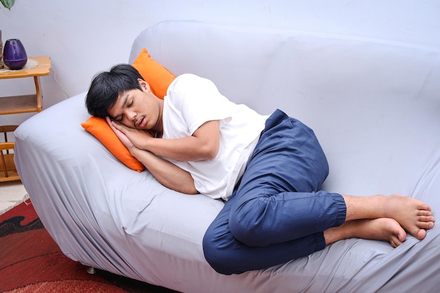 Exausto jovem asiático deitado no sofá confortável na sala de estar dormindo após um dia de trabalho duro