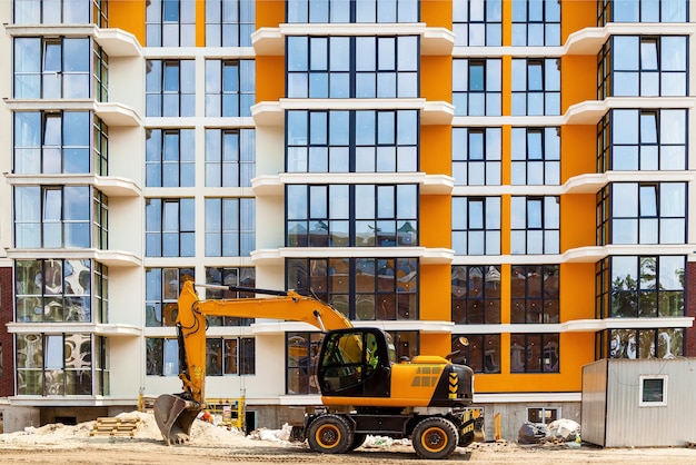 Una excavadora amarilla trabajando frente a un edificio residencial de gran altura con ventanas de vidrio.