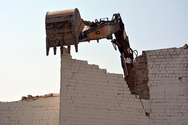 Una excavadora con un balde destruye una vieja pared de ladrillos para desmantelar una casa