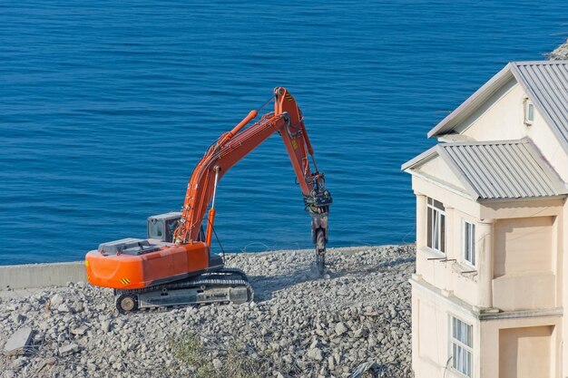 Excavadora con cizalla hidráulica contra un edificio demolido. desmantelamiento de construcción de emergencia a la orilla del mar.