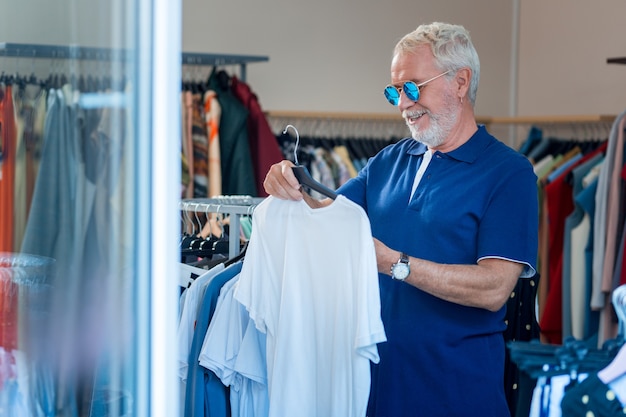 Excelente estilo. Cintura para arriba del hombre de moda de pelo gris con gafas de sol eligiendo un nuevo atuendo en la tienda de ropa mientras expresa deleite en su rostro