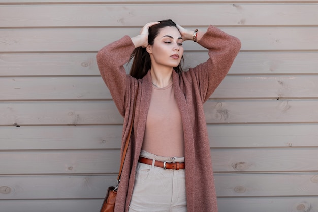 Foto excelente garota italiana em agasalhos da moda com casaco, pulôver e bolsa de couro posando perto de uma parede de madeira na rua