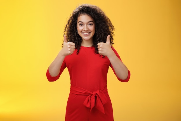 Excelente idea, buen trabajo. Retrato de una amiga encantadora encantadora y feliz de apoyo mostrando los pulgares para arriba como de pie en un vestido rojo sobre la pared amarilla sonriendo dando una respuesta positiva, gustando el concepto.