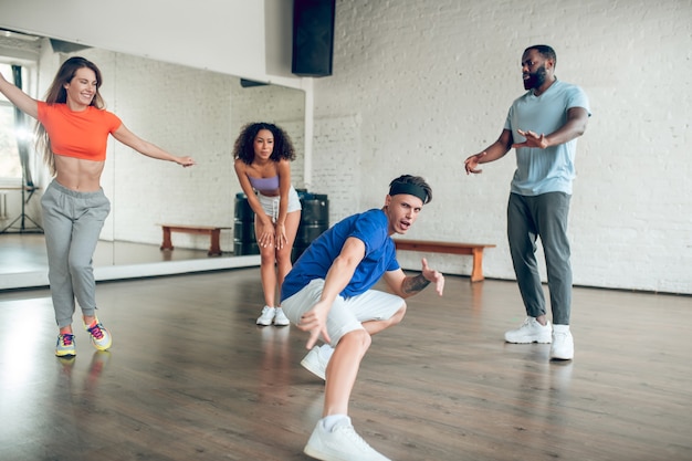 Excelente resultado. Joven chico emocional deportivo en diadema bailando en cuclillas estirando su pierna en círculo de amigos en el salón de baile