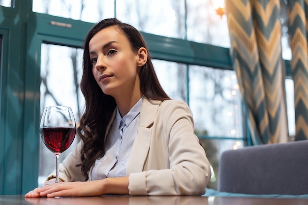 Excelente vinho. Mulher atraente reflexiva positiva tocando uma taça de vinho enquanto posa à mesa e descansa após o trabalho