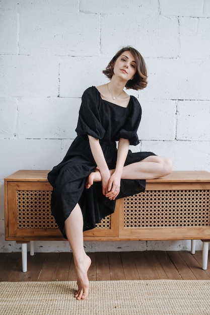 Foto excéntrica mujer laguida sentada en un armario bajo con un modesto vestido negro. con una pierna levantada. hermosa morena con pelo corto en el interior.