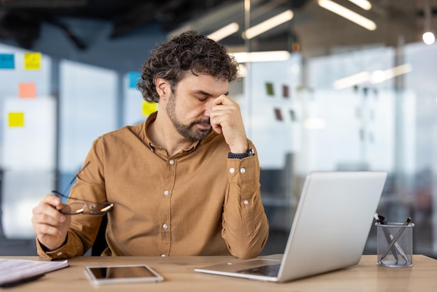 Excesivamente cansado y abrumado hombre de negocios tiene mareos y dolor en los ojos maduro trabajador masculino experimentado en el lugar de trabajo dentro de la oficina trabajando con computadora portátil