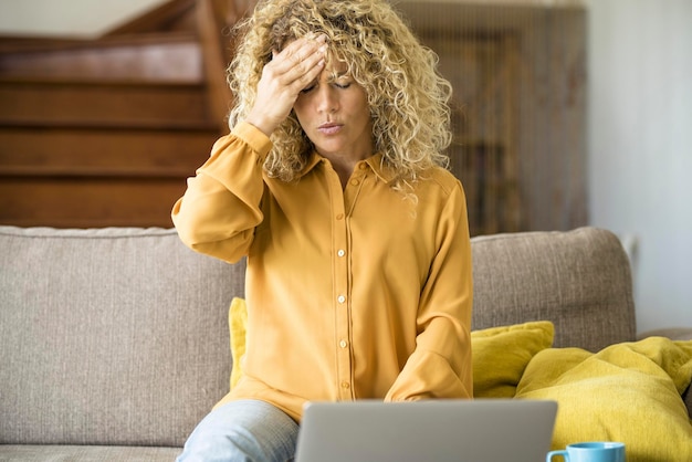 Exceso de trabajo y estrés malas condiciones de salud Mujer adulta tocando su frente y cabeza con dolor Mujeres cansadas y preocupadas usando computadora portátil en casa sentadas en el sofá Caída del mercado de valores