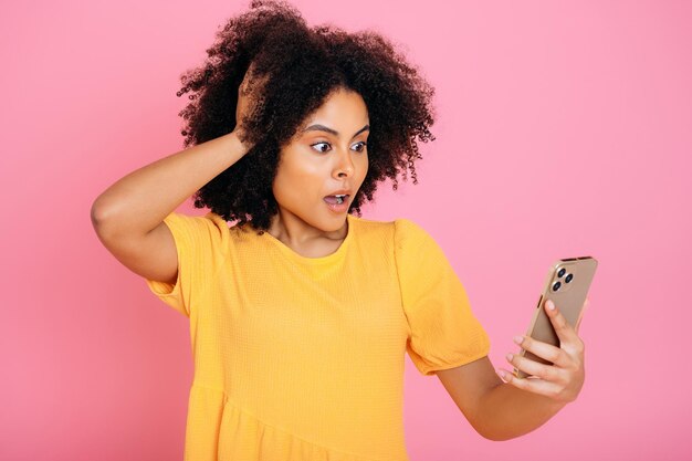 Foto excitada afro-americana ou brasileira mulher encaracolada olhando surpresa para seu smartphone segurando sua mão para sua cabeça recebeu uma notícia inesperada mensagem está perplexo isolado fundo rosa