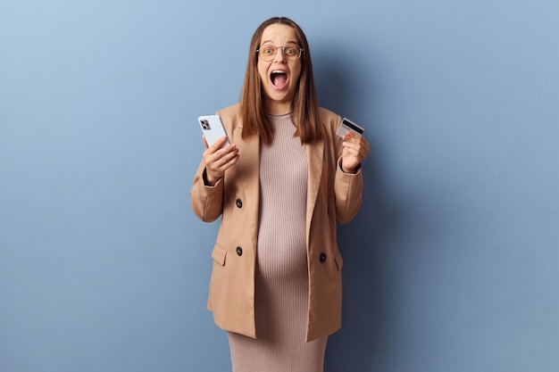 Foto excitada asombrada joven adulta embarazada con vestido y chaqueta posando aislada sobre un fondo azul haciendo pagos en línea disfrutando de compras en internet con teléfono inteligente y tarjeta de crédito