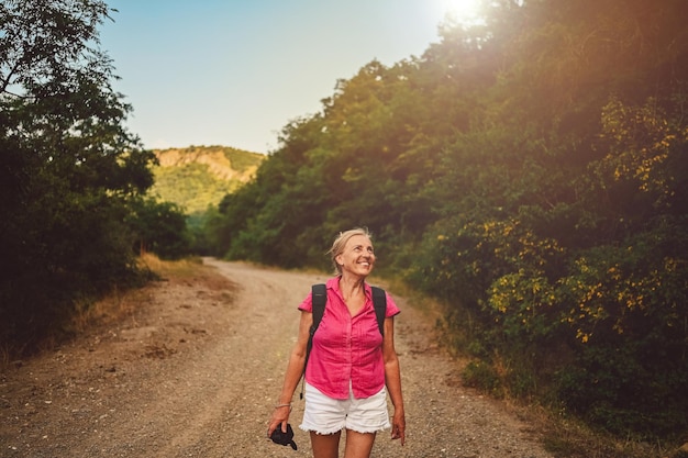 Foto excitada feliz mujer mayor mochilero turista caminando en el camino del bosque de verano al aire libre a la hora de la puesta del sol