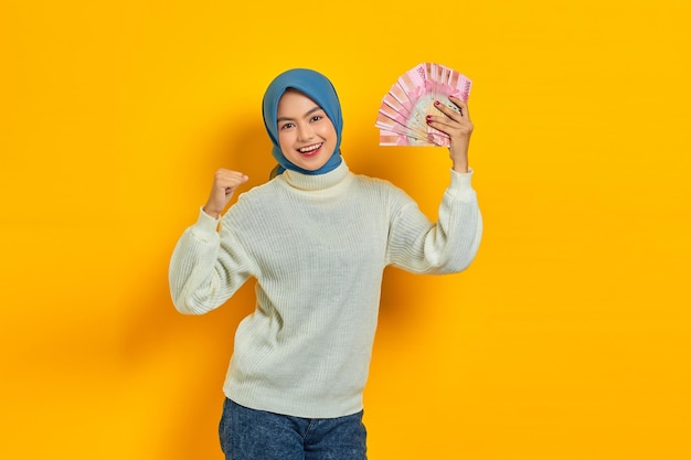 Excitada hermosa mujer musulmana asiática con suéter blanco sosteniendo dinero en efectivo en billetes de rupias indonesias y celebrando la suerte aislada sobre fondo amarillo Concepto de estilo de vida religioso de la gente