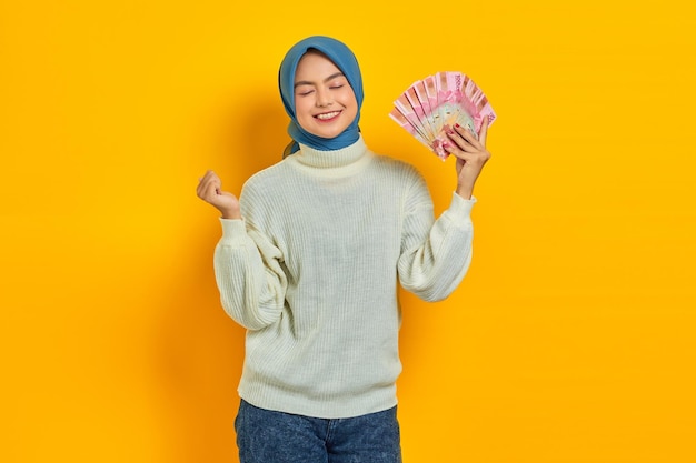 Excitada hermosa mujer musulmana asiática con suéter blanco sosteniendo dinero en efectivo en billetes de rupias indonesias y celebrando la suerte aislada sobre fondo amarillo Concepto de estilo de vida religioso de la gente