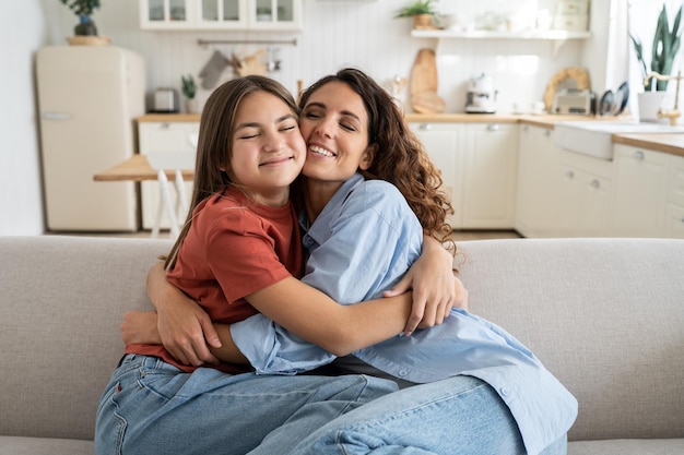 Foto excitada hija feliz abrazando a mamá con los ojos cerrados mientras están sentadas juntas en el sofá en casa