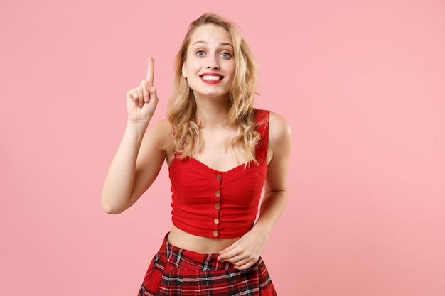 Excitada a una joven rubia con ropa sexy roja posando aislada en un retrato de estudio de fondo rosa pastel. Concepto de estilo de vida de las personas. Simulacros de espacio de copia. Mantenga el dedo índice hacia arriba con una gran idea nueva.