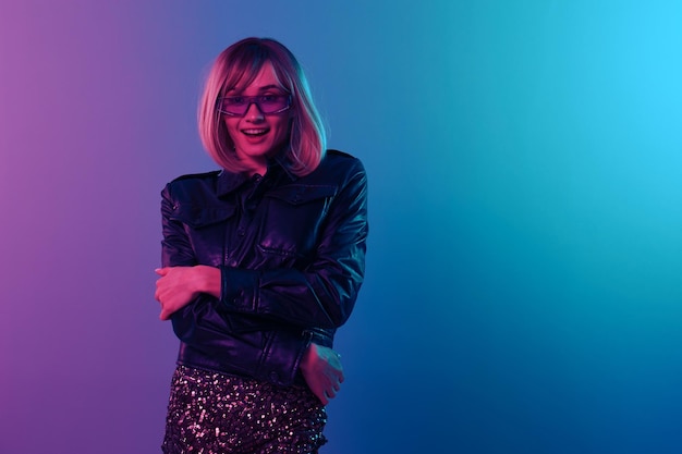 Excitada sexy hermosa mujer rubia en chaqueta de cuero vestido brillante gafas de sol de moda manos cruzadas posando aisladas en fondo de estudio de luz de color rosa azul fiesta de neón concepto Cyberpunk espacio de copia