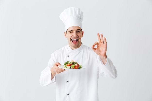 Excitado homem chef cozinheiro de uniforme, mostrando salada verde fresca em um prato isolado sobre uma parede branca, ok