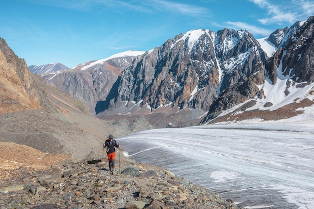 Excursionista con bastones de trekking cerca de una gran lengua glaciar en las altas montañas Paisaje alpino escénico con turistas cerca del glaciar a la luz del sol Hombre con gafas de sol y cámara entre montañas nevadas en un día soleado