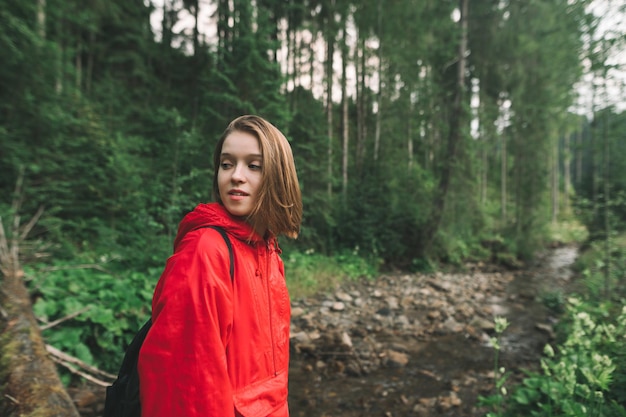 Excursionista de chica atractiva positiva en el bosque en el fondo de un arroyo de montaña y mira hacia otro lado