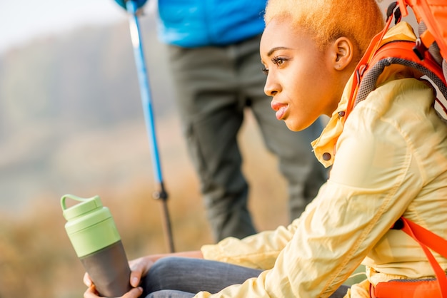 Excursionista de hermosa mujer africana en chaqueta amarilla bebiendo agua de la botella sentado al aire libre