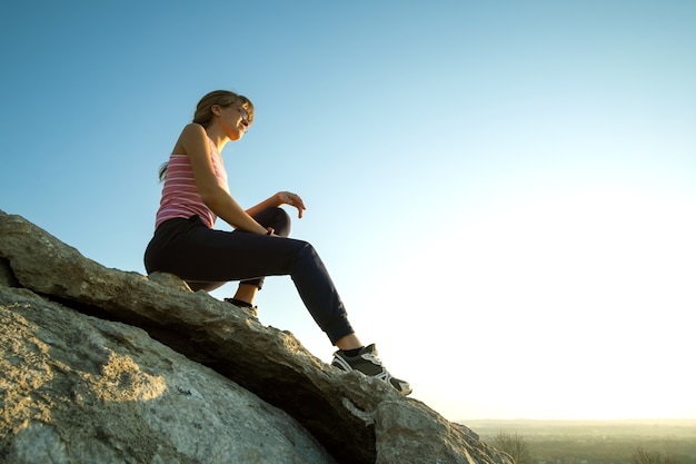 Excursionista mujer sentada sobre una gran roca empinada disfrutando de un cálido día de verano. Escalador joven descansando durante la actividad deportiva en la naturaleza. Recreación activa en concepto de naturaleza.