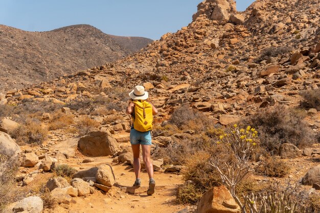 Un excursionista con sombrero y mochila amarilla caminando por el sendero del cañón hacia el Mirador de las Peñitas, Fuerteventura, Islas Canarias. España