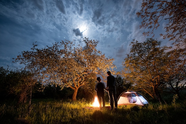 Excursionistas cerca de fogata y carpa turística en la noche.