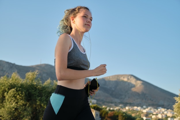 Executando uma jovem adolescente em fones de ouvido de roupas de fitness com smartphone