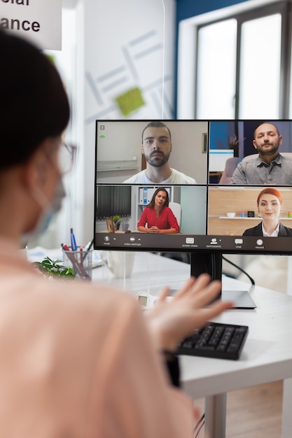 Executive Manager mit medizinischer Gesichtsmaske, die Managementstatistiken mit dem Remote-Team diskutiert, das eine Online-Videocall-Meeting-Konferenz auf einem Laptop hat, der im Startup-Büro arbeitet. Telefonkonferenz auf dem Bildschirm