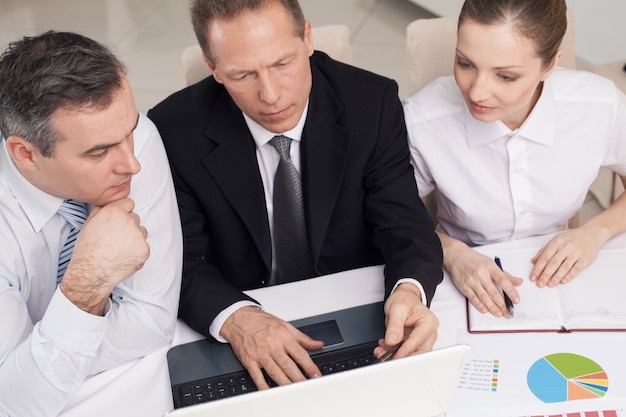 Foto executivos no trabalho. vista superior de três alegres executivos em trajes formais, sentados à mesa e olhando para o laptop